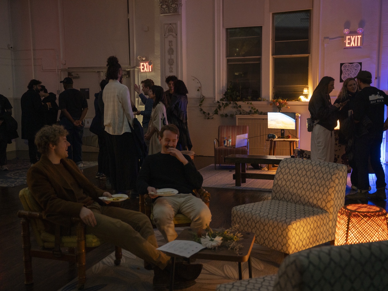 Two attendees of the Brooklyn local AI meetup laughing on a striped armchair in the lounge, cool ambient light, an EXIT sign in the background. The Grove, Greenpoint, NYC.